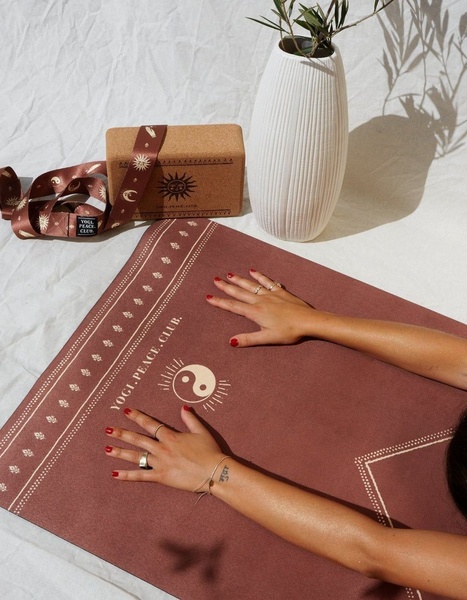 A high-angle product shot of a brown yoga mat and matching cork block, featuring a person's hands resting on the mat surface in a calming studio environment.